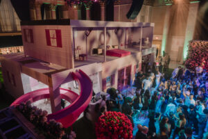 Two-story Barbie Dream House installation at a corporate event, surrounded by attendees, showcasing vibrant lighting and floral decorations.