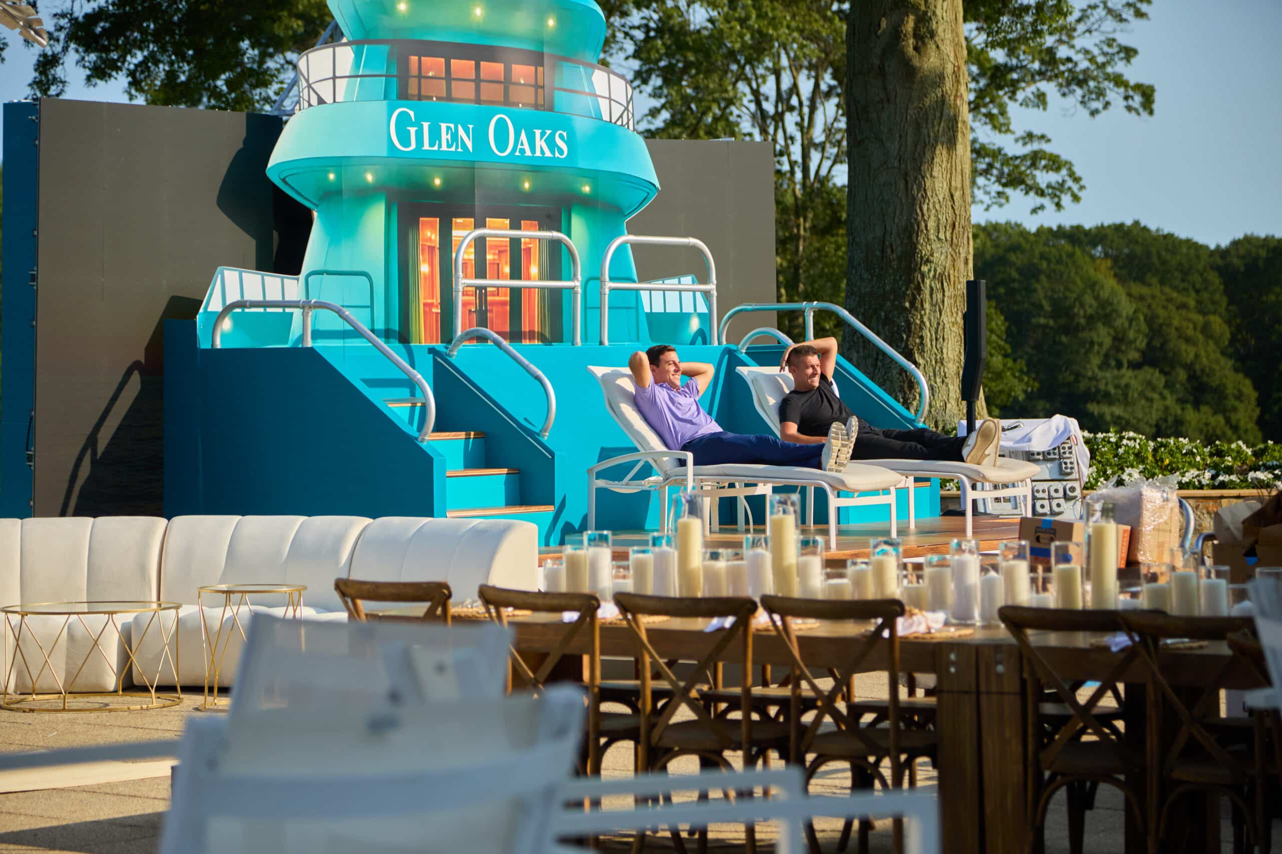 Two men relaxing on lounge chairs in front of a vibrant blue themed setup with "Glen Oaks" signage, surrounded by tables and decor for a luxury tropical event at Glen Oaks Country Club.